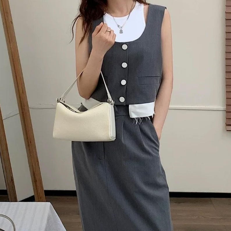 May include: A cream-colored, rectangular handbag with a single shoulder strap. The bag is held in front of a person wearing a gray vest with white trim and a matching skirt. The vest has white buttons and a pocket.