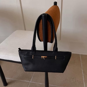 May include: A black handbag with a rectangular shape and two handles. The bag features gold-colored hardware, including a clasp and decorative accents. The bag is hanging on a chair.