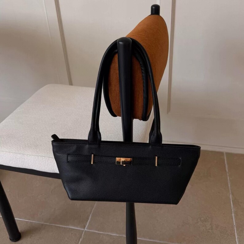 May include: A black handbag with a rectangular shape and two handles. The bag features gold-colored hardware, including a clasp and decorative accents. The bag is hanging on a chair.