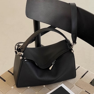 May include: Black leather shoulder bag with a top handle and a detachable shoulder strap. The bag has a zippered closure and silver-tone hardware. The bag is sitting on a chair.