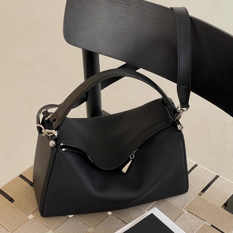 May include: Black leather shoulder bag with a top handle and a detachable shoulder strap. The bag has a zippered closure and silver-tone hardware. The bag is sitting on a chair.