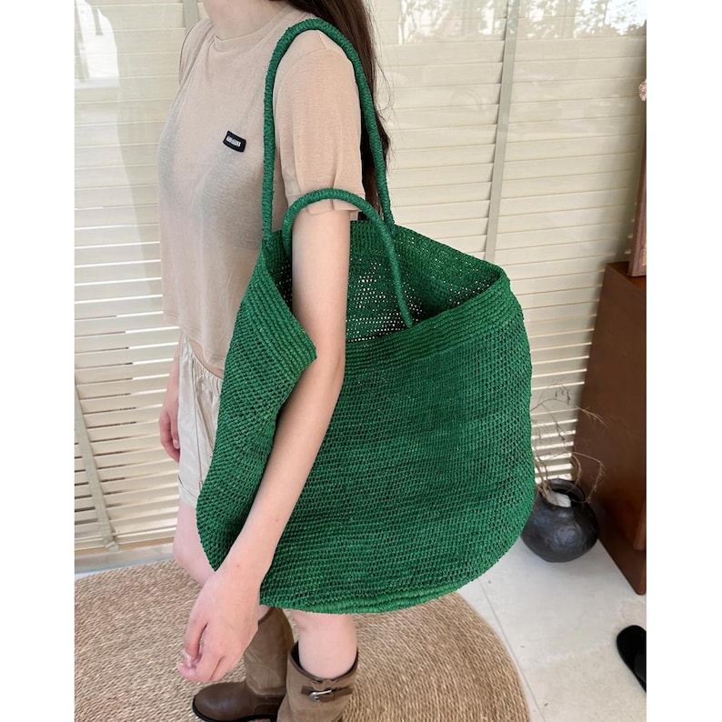 May include: A large, deep green woven tote bag featuring a textured, straw-like material construction with two long, integrated shoulder straps. The bag has a spacious, open-top design and a consistent, uniform dark green color throughout the exterior.