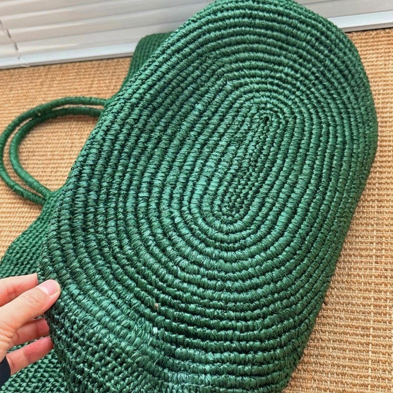 May include: A deep forest green tote bag constructed from tightly coiled, woven material. The bag features a distinct oval-shaped base with a continuous spiral pattern and two matching green woven handles attached to the side of the main body.