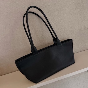 May include: A black rectangular tote bag with two long, slender handles. The bag is made of a smooth material and has a simple, minimalist design. The handles are attached to the top of the bag and curve upwards.
