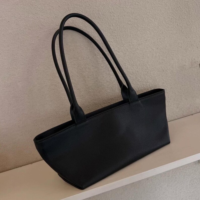 May include: A black rectangular tote bag with two long, slender handles. The bag is made of a smooth material and has a simple, minimalist design. The handles are attached to the top of the bag and curve upwards.