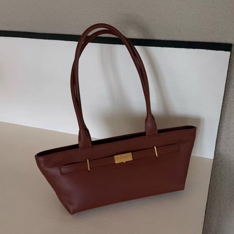 May include: A brown leather tote bag with long handles. The bag features a rectangular shape with a gold-colored clasp and accents. The bag is designed with a simple, elegant style.