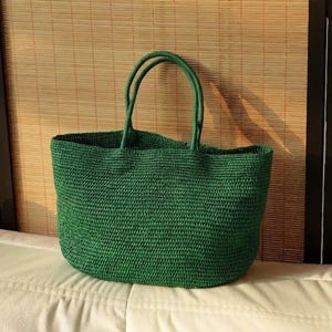 May include: A green woven tote bag with two handles. The bag is made of a textured material and has a rectangular shape. The bag is sitting on a white surface with a bamboo blind in the background.