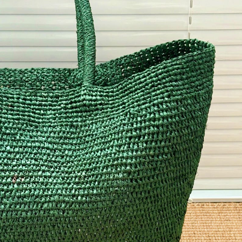May include: A deep forest green tote bag constructed from a woven, textured material. The bag features a wide, open top design and a sturdy, integrated handle that extends upward from the main body, showcasing a consistent, dense weave pattern.