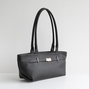 May include: Black tote bag with long handles. The bag features a rectangular shape with a textured surface and a gold-colored clasp closure. The handles are thin and attached to the top of the bag.