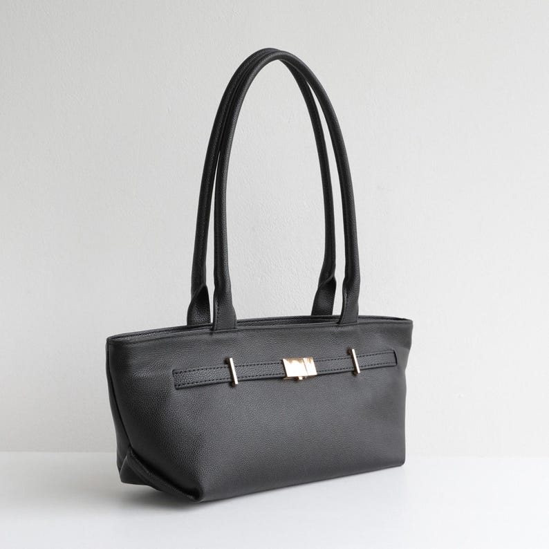 May include: Black tote bag with long handles. The bag features a rectangular shape with a textured surface and a gold-colored clasp closure. The handles are thin and attached to the top of the bag.