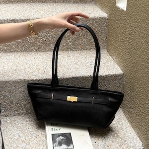 May include: Black leather tote bag with a rectangular shape and two top handles. The bag features a gold-colored clasp and accents. The bag is held by a person. A book with the text "for Her" is visible.