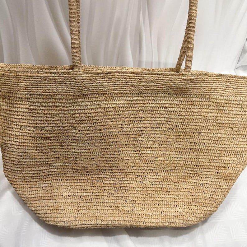 May include: A large, tan-colored woven straw tote bag featuring a wide, rectangular body with a rounded bottom edge and two long, sturdy shoulder straps attached to the top rim. The texture is consistent throughout the entire surface.
