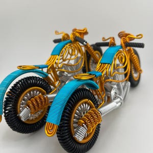 May include: Two gold wire sculptures of motorcycles with blue accents. The motorcycles are facing the same direction, with the front motorcycle partially obscured by the rear motorcycle.