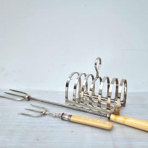 May include: A silver-plated toast rack with a handle, and two serving forks with cream-coloured handles. The forks have a silver-plated metal design. The items are arranged on a light-coloured surface.