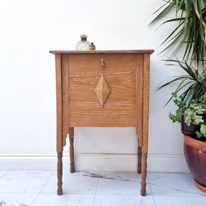 May include: A light brown wooden side table featuring a rectangular top, four slender turned legs, and a front panel decorated with a central diamond-shaped wooden inlay. A small metal knob is positioned above the diamond detail on the front.