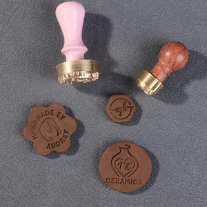 May include: Two wax seal stamps with brass bases and wooden handles, one pink and one brown. Four brown wax seals are also shown, with various designs and text, including "handmade by Audrey" and "TK Ceramics".