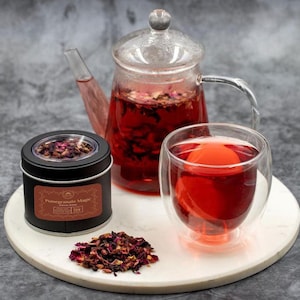 May include: A glass teapot and a double-walled glass of red tea sit on a white tray, alongside a black tin of tea and a small pile of loose tea leaves. The tea is a vibrant red color, and the scene is set against a gray background.