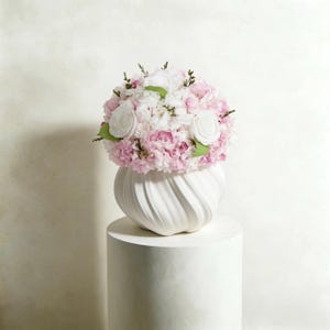 以下が含まれることがあります： 白い渦巻き模様のセラミック花瓶に、ピンクと白の花のブーケが入っています。アレンジメントにはバラとアジサイが含まれ、緑色のアクセントがあります。花瓶は白い円筒形の台座の上に置かれています。