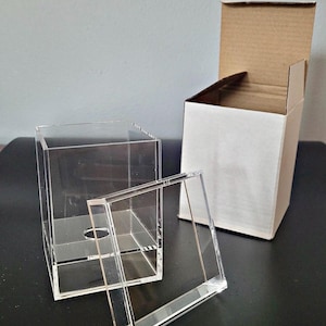 Puede incluir: Una caja de exhibición de acrílico transparente con una tapa extraíble. La caja está vacía y tiene un pequeño agujero en la parte inferior. La caja está sentada en una mesa junto a una caja de cartón.