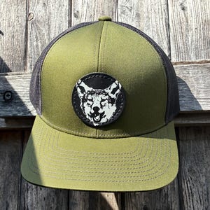Puede incluir: Gorra de camionero verde oliva con parte trasera de malla negra y visera curva. La gorra presenta un parche circular negro con una ilustración blanca de la cabeza de un lobo. Sobre un fondo de madera envejecida.