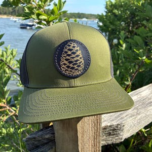 May include: Olive green trucker hat with a black mesh back and a pine cone patch. The patch is a dark blue circle with a gold pine cone design. The hat is sitting on a wooden post with a scenic background of water and trees.