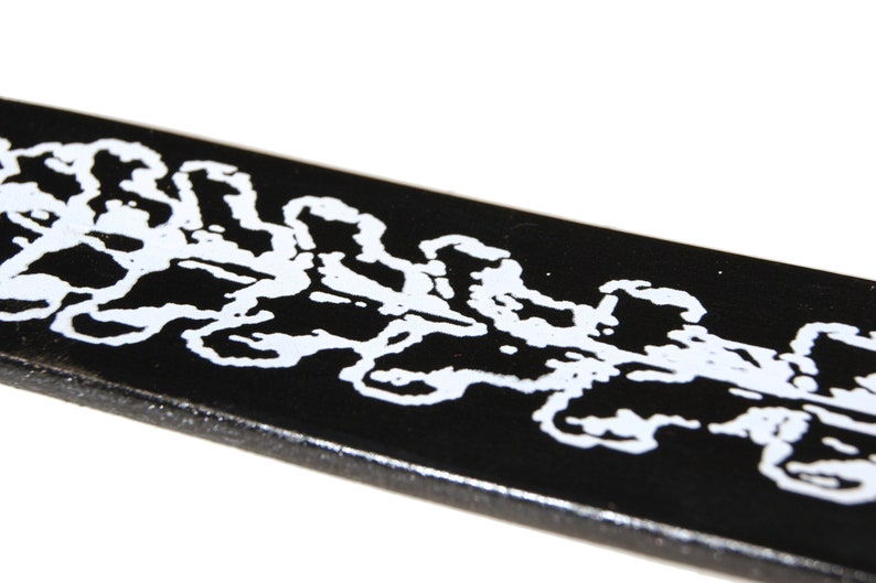Op de afbeelding: Zwarte leren riem met een wit ontwerp van een herhalend patroon van bladeren.
