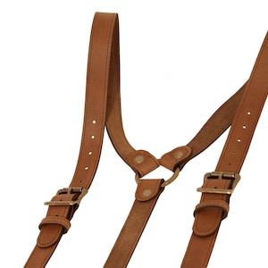 Puede incluir: Tirantes de cuero marrón con hebillas de latón y un anillo de metal. Los tirantes tienen correas ajustables y están diseñados para sujetar los pantalones.