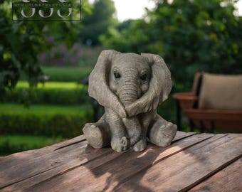 Puede incluir: Una figura de elefante gris con la trompa enrollada, sentada sobre una mesa de madera. El elefante tiene orejas grandes y una textura detallada. El fondo es una escena exterior borrosa con vegetación.