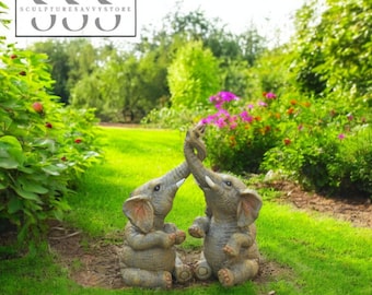 May include: Two gray elephant statues with trunks intertwined, sitting on a grassy lawn. The statues have detailed features and are surrounded by green foliage and colorful flowers. The logo "Sculptures Savvy Store" is in the top left corner.