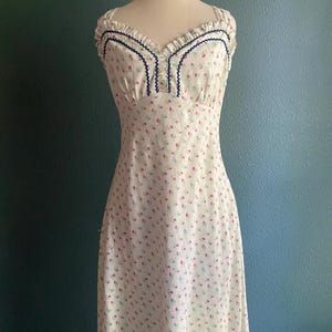 May include: A vintage-style, floor-length dress on a mannequin. The dress is cream-coloured with a delicate floral pattern in pink and blue. It features a sweetheart neckline with blue trim and ruffled edges.
