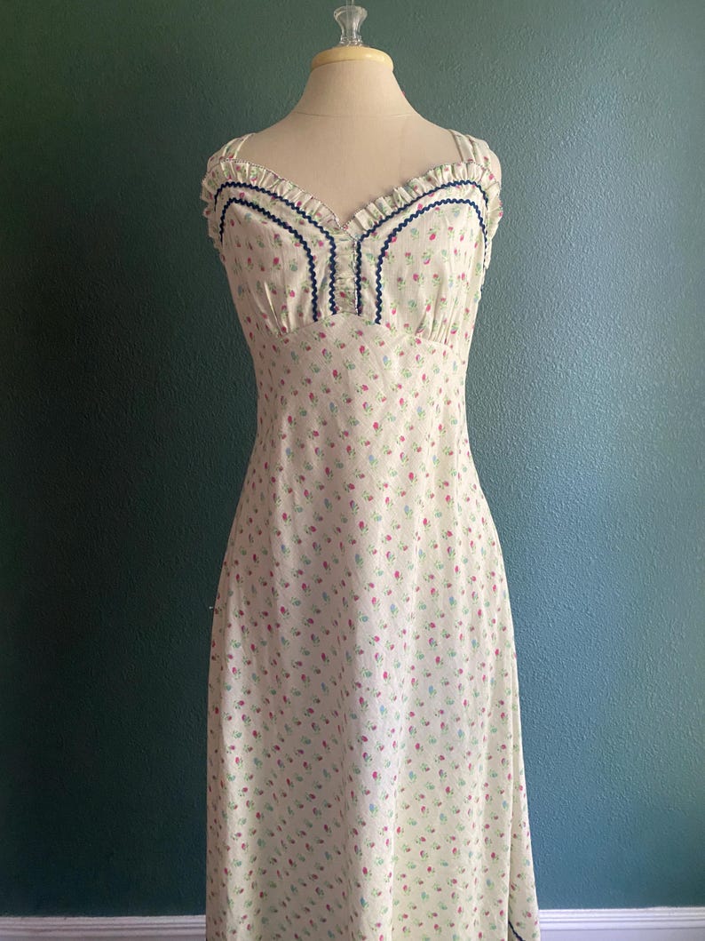 May include: A vintage-style, floor-length dress on a mannequin. The dress is cream-coloured with a delicate floral pattern in pink and blue. It features a sweetheart neckline with blue trim and ruffled edges.