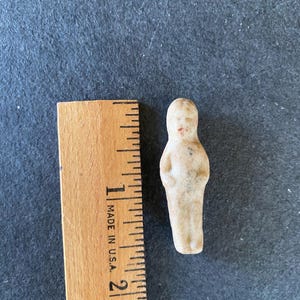 May include: A small, off-white doll figurine with simple features, including a red dot for a mouth. The doll is approximately 1.5 inches tall. A wooden ruler with black markings and the text "MADE IN U.S.A." is next to the doll.