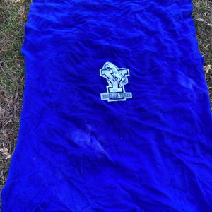 May include: A vibrant blue blanket with a fringed edge. The blanket features a white and blue emblem with the text "BRIGHAM YOUNG UNIVERSITY". The blanket is spread out on a bed of grass.