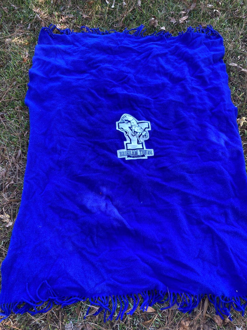May include: A vibrant blue blanket with a fringed edge. The blanket features a white and blue emblem with the text "BRIGHAM YOUNG UNIVERSITY". The blanket is spread out on a bed of grass.