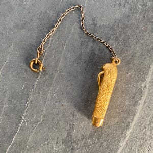 May include: A gold-toned metal pocket knife pendant featuring an intricate, etched leaf-like pattern on its surface. The small, closed knife is attached to a delicate matching metal chain that includes a circular spring ring clasp at the end.