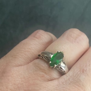 May include: A silver ring featuring a marquise-cut, vibrant green gemstone. The ring's band has an ornate, openwork design. The gemstone is the focal point, set prominently in the center of the ring.