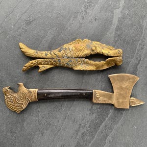 May include: A brass nutcracker shaped like a fish with textured scales and a brass tool featuring an axe head on one end, a bottle opener shaped like a stylized animal head on the other, and a dark brown wooden handle with the word Thailand.