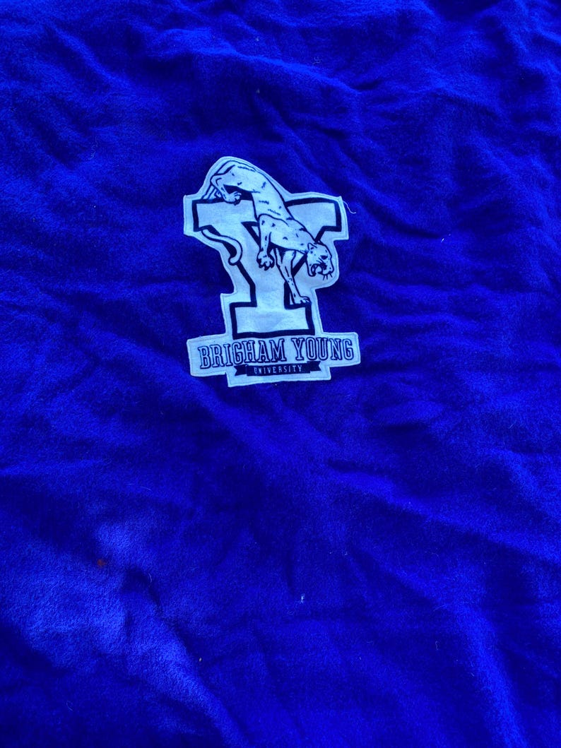 May include: Royal blue fabric with a white patch featuring a cougar and the text "BRIGHAM YOUNG UNIVERSITY". The cougar is in a leaping pose. The fabric has a soft, textured appearance.