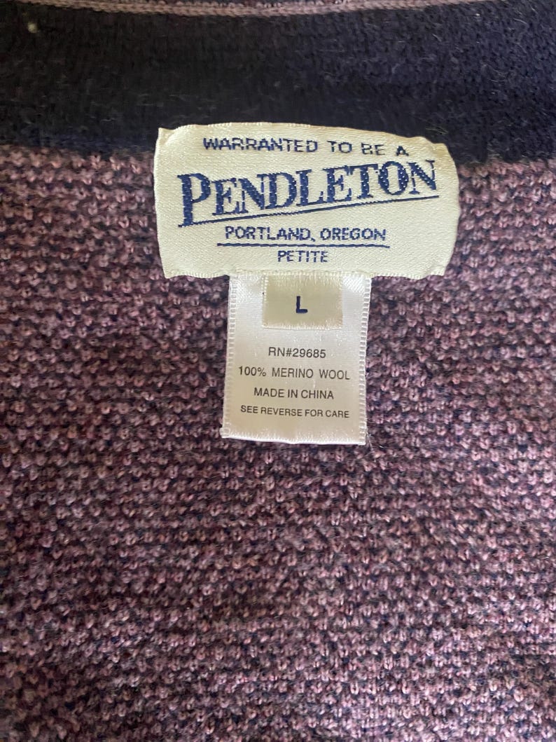 Puede incluir: Primer plano de un su&eacute;ter Pendleton morado y azul marino. La etiqueta dice "PENDLETON PORTLAND, OREGON PETITE" e incluye la talla "L". La etiqueta tambi&eacute;n indica que el su&eacute;ter es 100% lana merino y est&aacute; hecho en China.