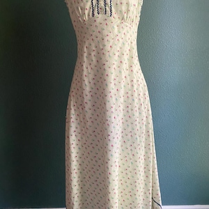 May include: A vintage-style, floor-length dress with a light yellow background and a delicate floral pattern in red and blue. The dress features a fitted bodice with a ruffled neckline and navy blue trim. The hemline also has navy blue trim.