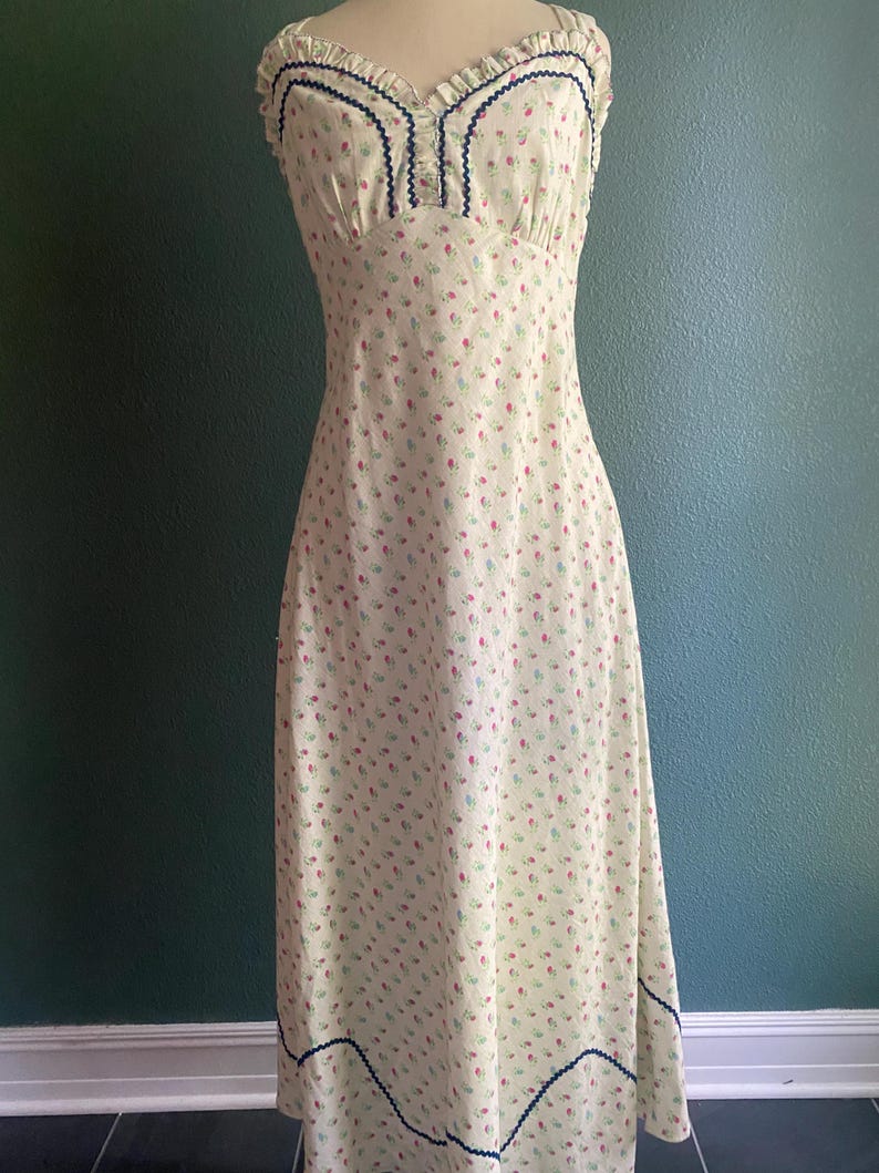 May include: A vintage-style, floor-length dress with a light yellow background and a delicate floral pattern in red and blue. The dress features a fitted bodice with a ruffled neckline and navy blue trim. The hemline also has navy blue trim.
