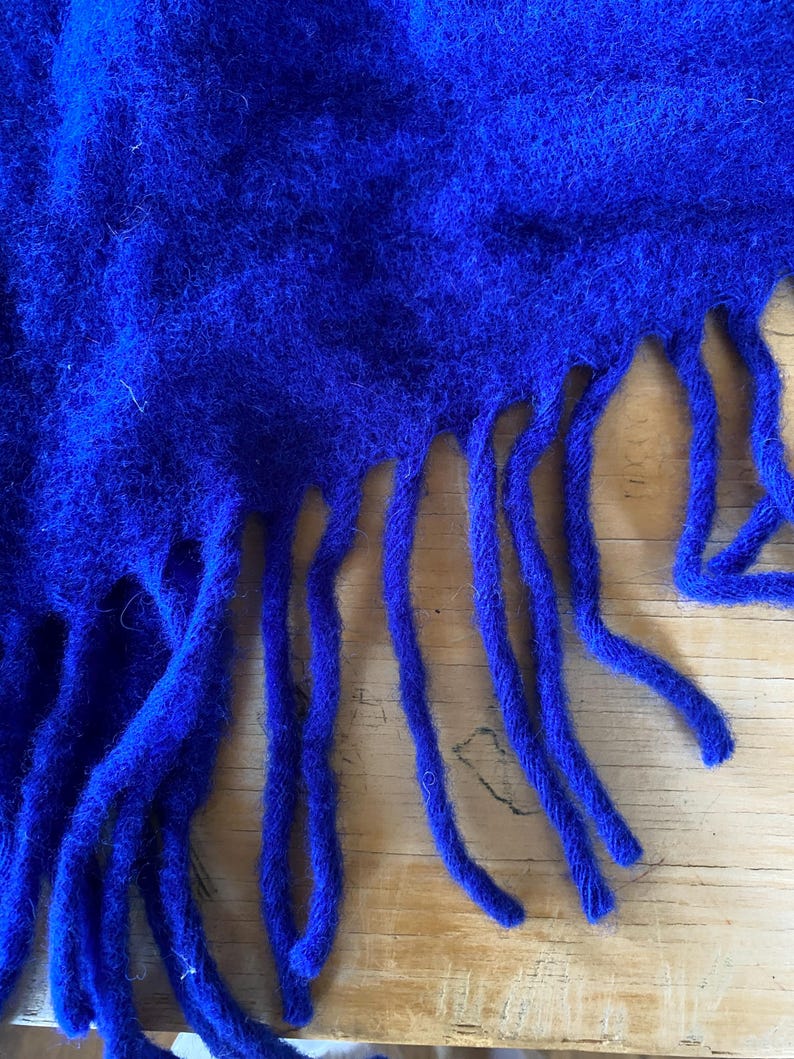 May include: Close-up of a vibrant blue scarf with a textured, fuzzy appearance. The scarf has long, thick fringe along the bottom edge. The fabric appears soft and warm, suggesting a cozy accessory.