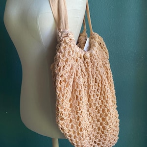 May include: A peach-colored crocheted tote bag with matching straps is displayed on a mannequin. The bag has a net-like design and a gathered top. The straps are made of a soft, fabric material. The bag is a light, neutral color.