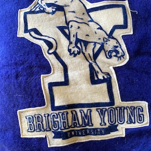 May include: A blue felt patch featuring a cream-colored cougar and the text "BRIGHAM YOUNG UNIVERSITY." The cougar is leaping over a large letter "Y." The text is in a dark blue color.