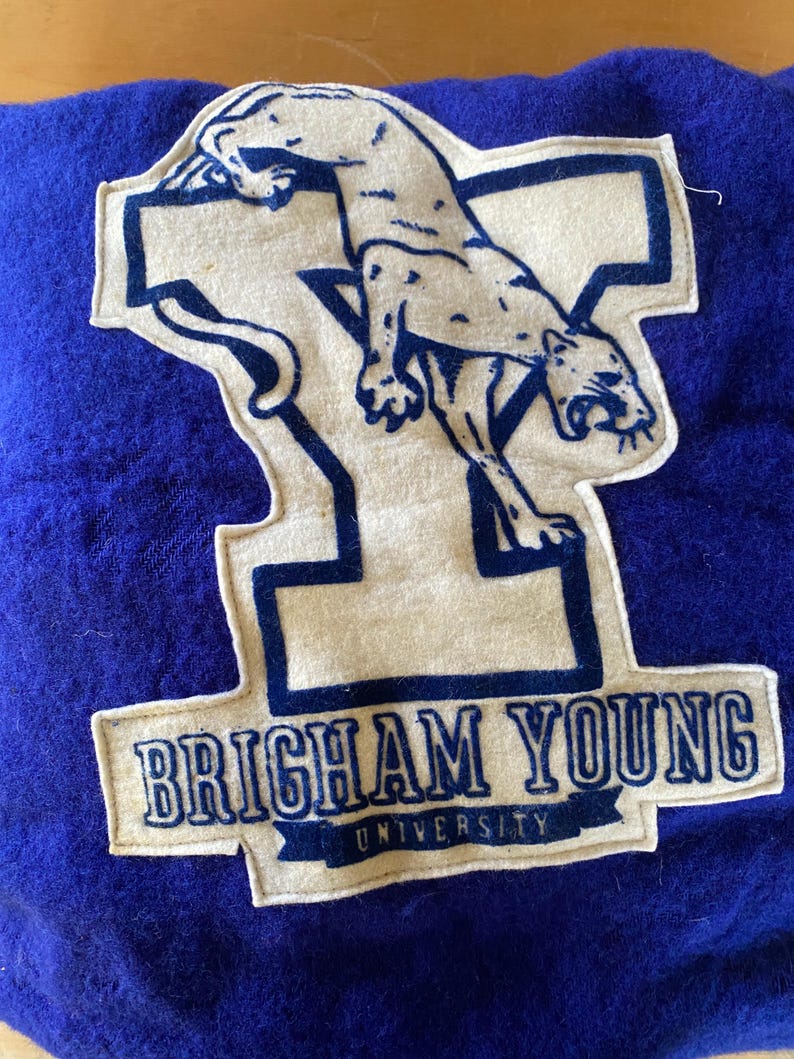 May include: A blue felt patch featuring a cream-colored cougar and the text "BRIGHAM YOUNG UNIVERSITY." The cougar is leaping over a large letter "Y." The text is in a dark blue color.
