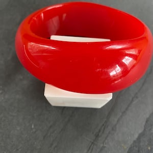 May include: A vibrant red bangle bracelet. The smooth, glossy surface reflects light, highlighting its rounded shape. The bracelet is resting on a white rectangular block, set against a dark background. This bold accessory is a statement piece.