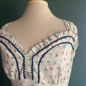 May include: A cream-coloured dress with a floral pattern of pink and blue flowers. The dress features a ruffled neckline and straps, with navy blue trim. The dress is displayed on a mannequin.