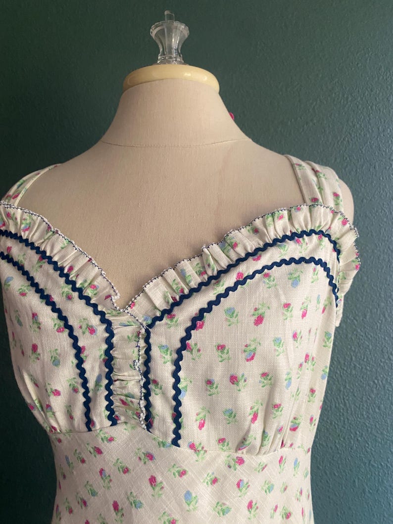 May include: A cream-coloured dress with a floral pattern of pink and blue flowers. The dress features a ruffled neckline and straps, with navy blue trim. The dress is displayed on a mannequin.