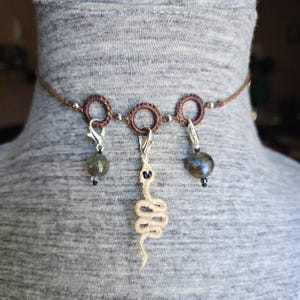 Pode incluir: Um colar gargantilha de macramé castanho com três anéis circulares. Dois fechos de mosquetão prateados seguram contas redondas cinzentas iridescentes, enquanto o anel central contém um pingente de cobra bege claro feito de corda.
