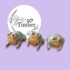 May include: Three ceramic frog figurines with blue and green glaze, yellow bellies, and large black eyes. The frogs are arranged in a row on a purple background.